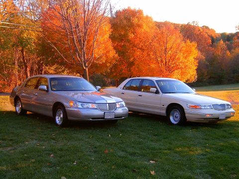 1996 Mercury Grand Marquis - Two silver full-size sedans parked side by side on grass in front of vivid orange autumn foliage; the left car is a Lincoln Town Car and th…