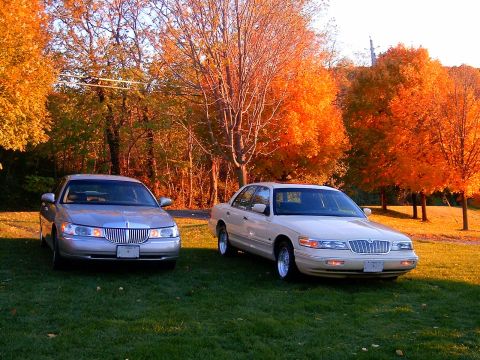 1996 Mercury Grand Marquis - Two full-size sedans parked side by side on grass in front of brilliant orange fall foliage: a silver Lincoln Town Car on the left and a cr…