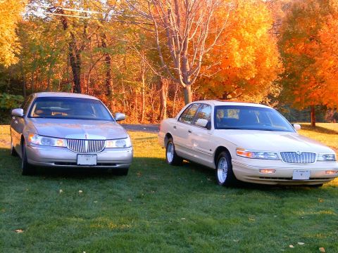 1996 Mercury Grand Marquis - Two full-size sedans parked side-by-side on grass in front of autumn-colored trees; a silver Lincoln Town Car on the left and a cream/tan M…