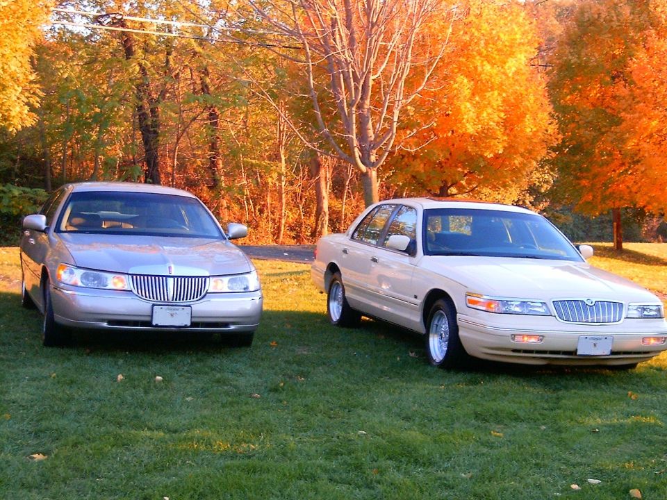 Silver Lincoln and tan Mercury sedans parked on grass with fall foliage behind.