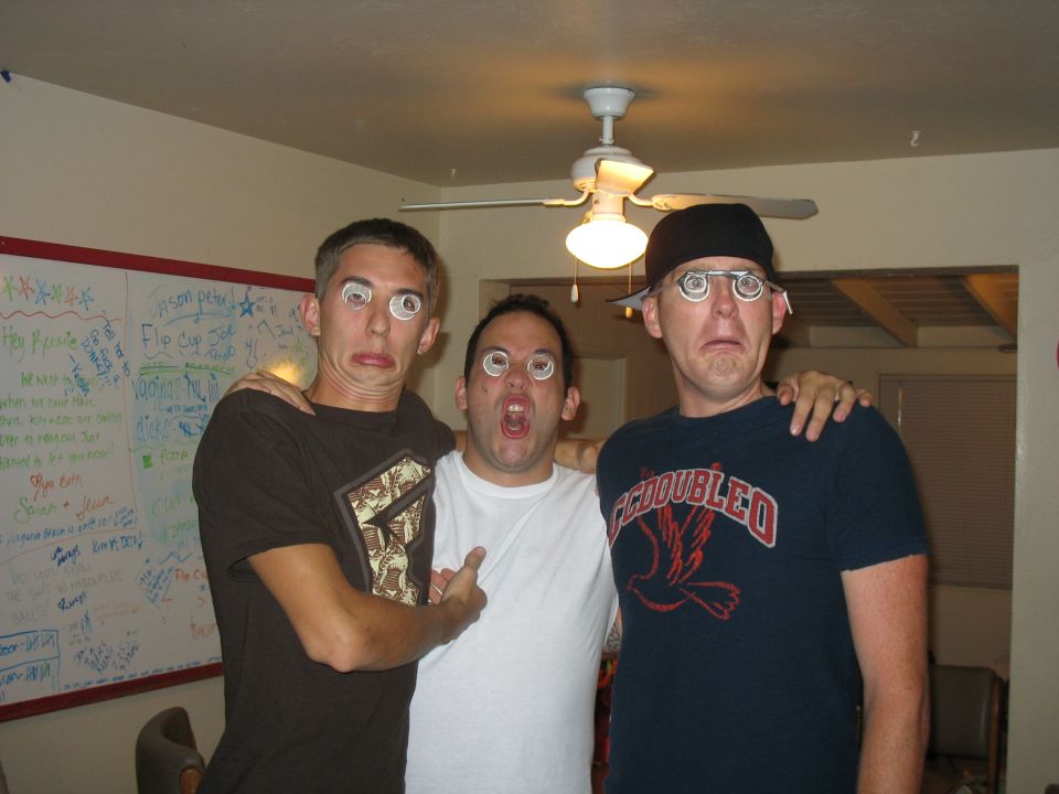 Three men making silly faces with eyes-painted glasses indoors under a ceiling fan