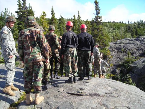 2003 Jeep TJ - A group of soldiers stand together on a rocky terrain, surrounded by trees under a bright sky.