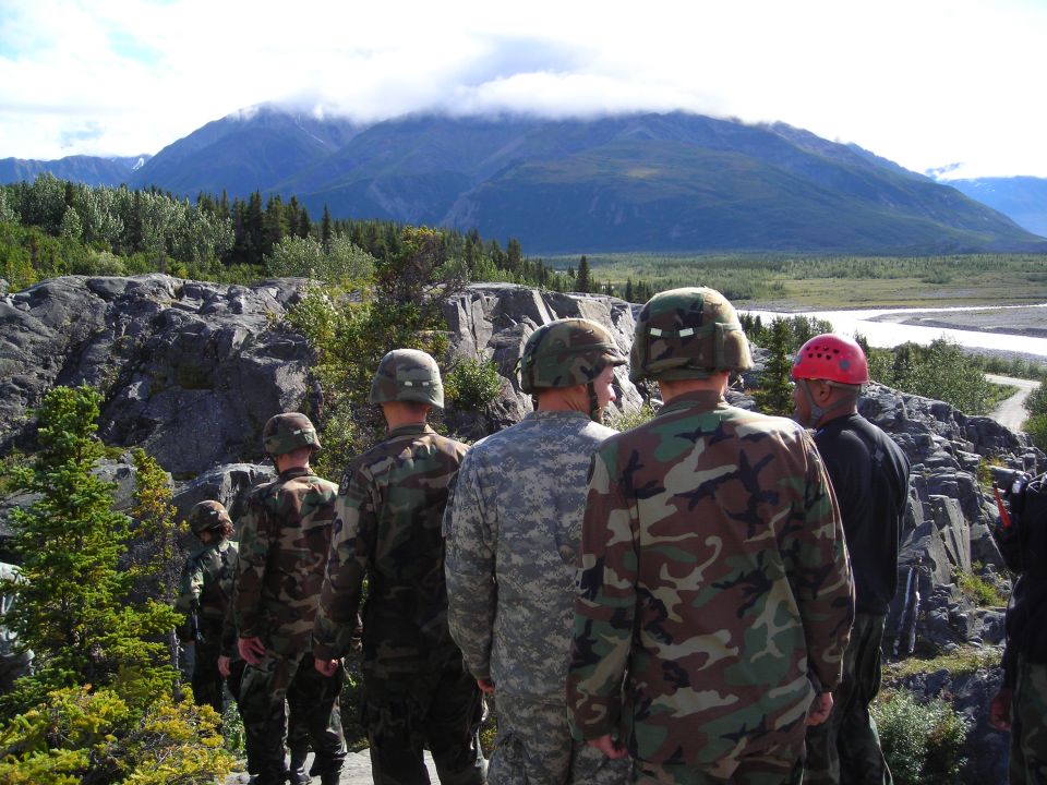 Soldiers stand on rocky terrain overlooking a wide river valley with mountains in the background