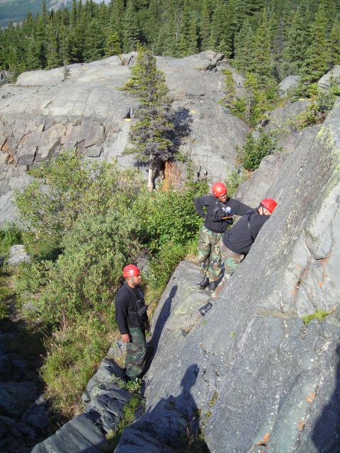 2003 Jeep TJ - Three individuals wearing helmets and camouflage pants are engaged in rock climbing on a steep, cliffside amidst a forested mountain landsc…