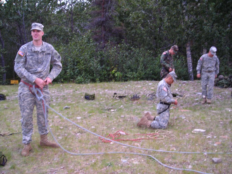 Soldiers in camouflage uniforms preparing ropes outdoors with trees in the background