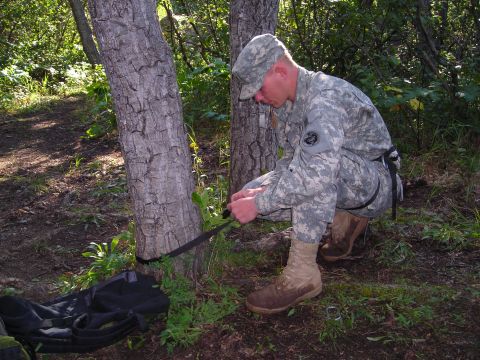 2003 Jeep TJ - A soldier crouches to secure a strap around a tree in a forest setting, with a backpack on the ground nearby.