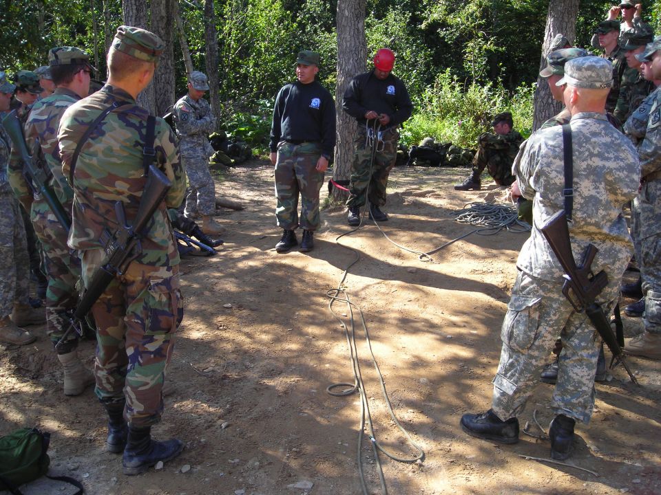 Military personnel in camouflage uniforms stand in a dirt clearing surrounded by trees, with ropes on the ground