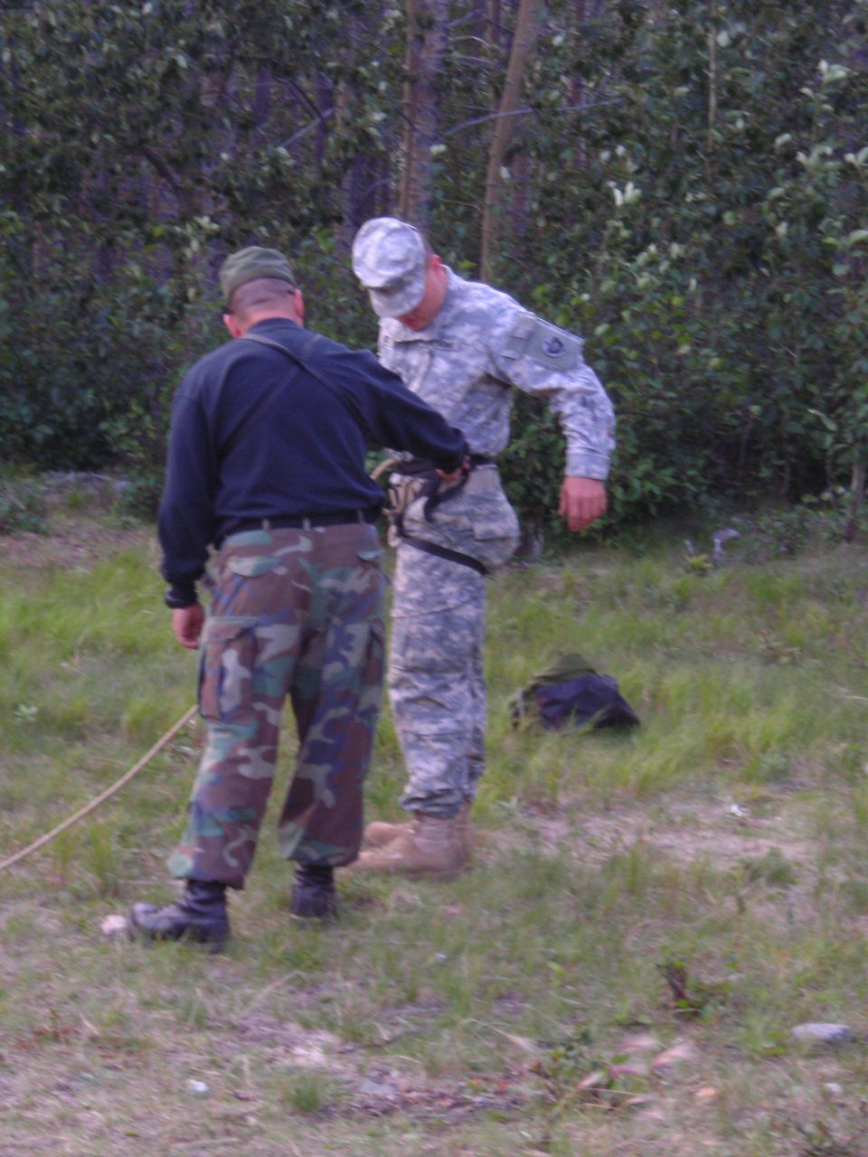 Two men in military uniforms, one adjusting gear on the other, outdoors