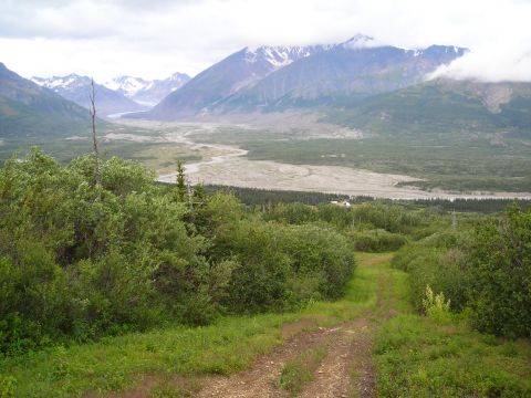 2003 Jeep TJ - A dirt track leads through lush vegetation towards a vast valley, framed by imposing mountains under a cloudy sky.