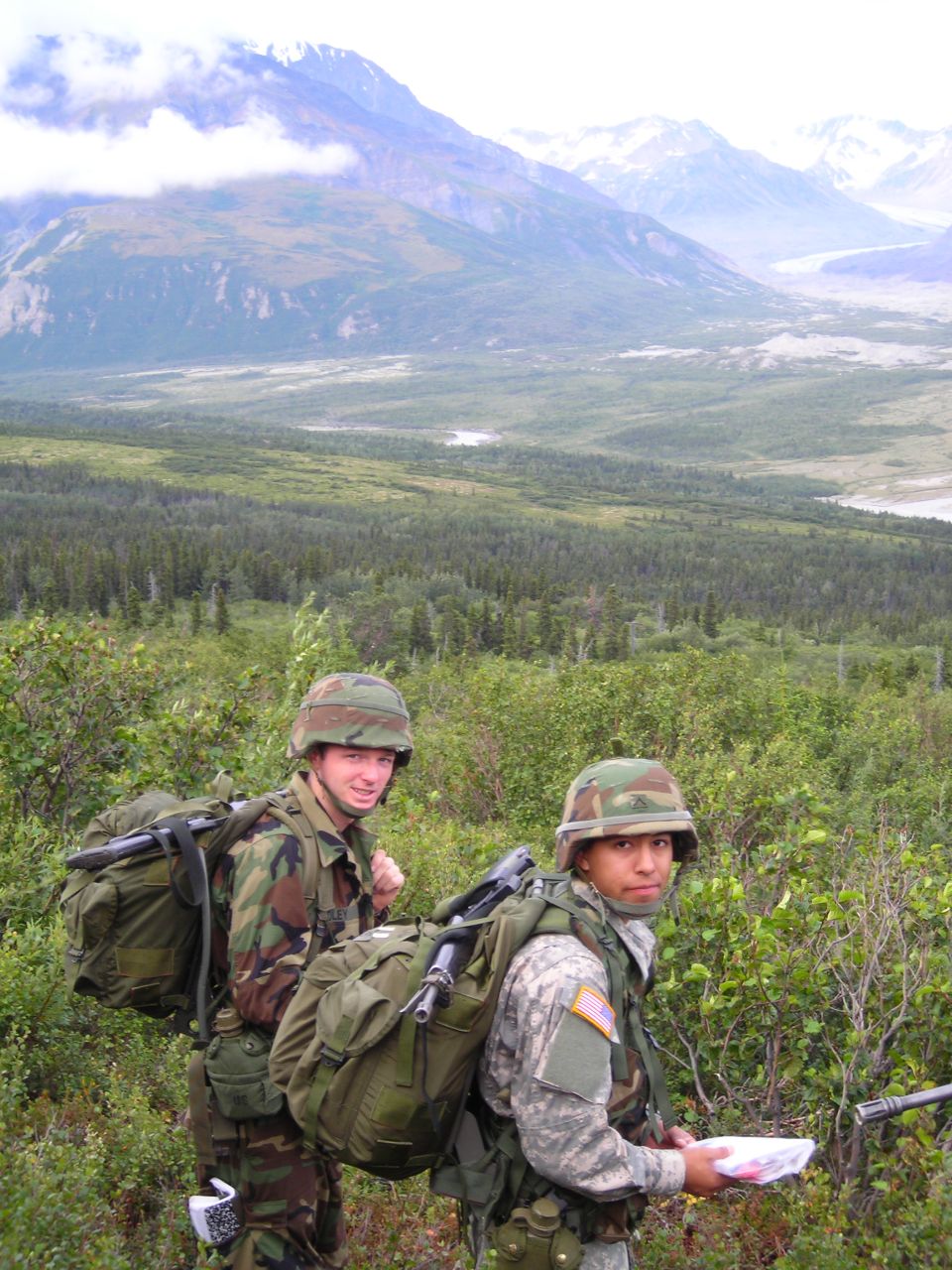 Two soldiers wearing helmets and carrying large backpacks walk through a wooded area with mountains in the background
