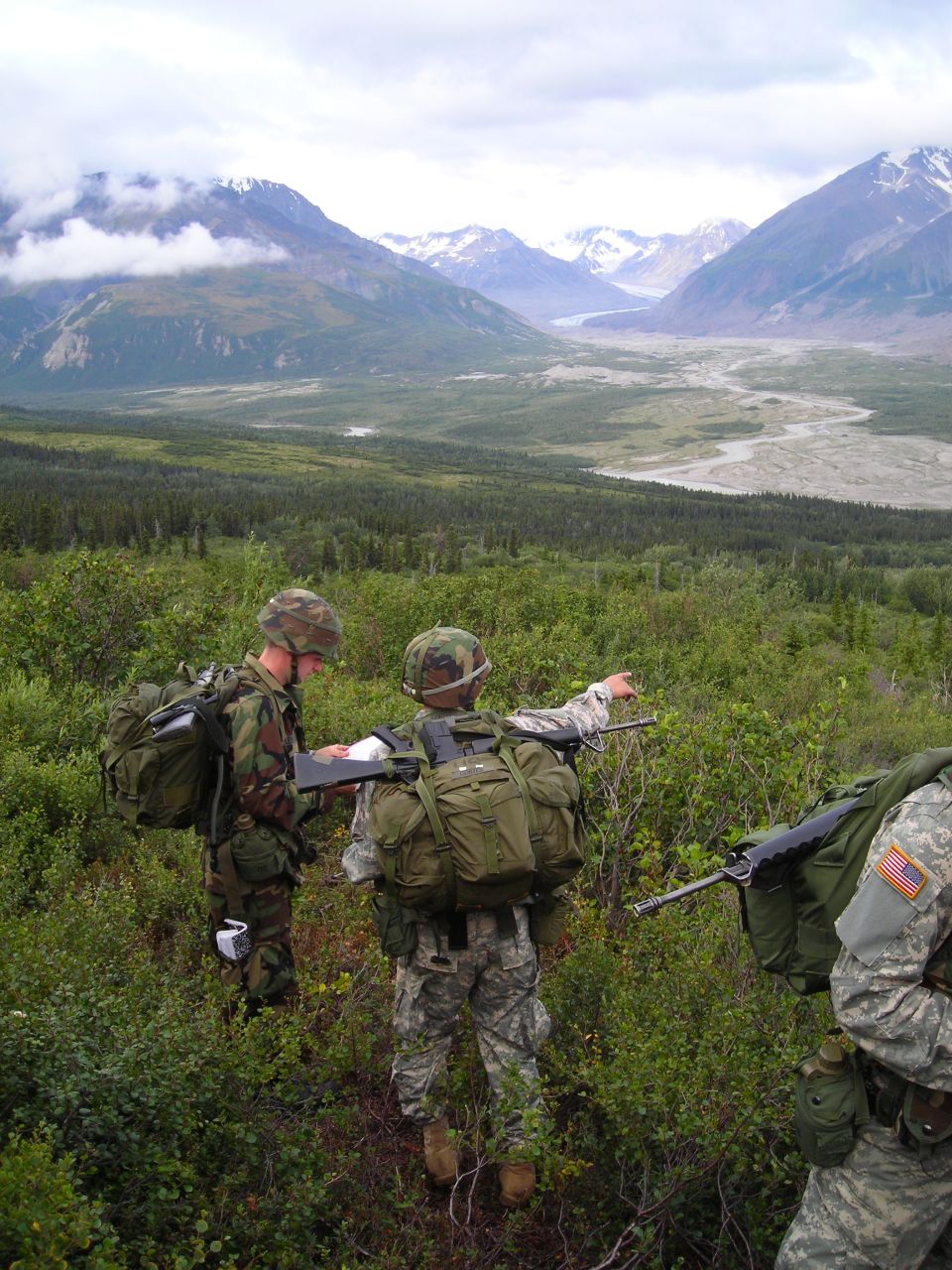 Soldiers with backpacks and rifles in a green, mountainous terrain with a river valley