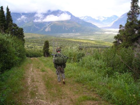 2003 Jeep TJ - A soldier with a large backpack walks down a dirt path surrounded by trees, with a vast mountainous landscape stretching out before them.
