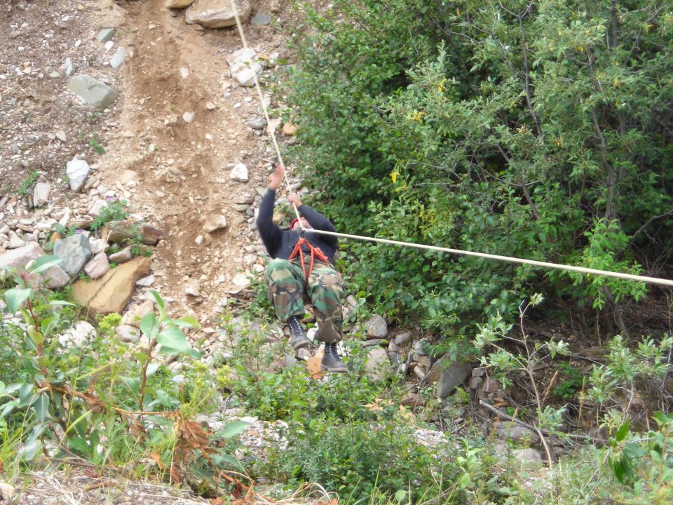 Person in camouflage pants and dark shirt rappelling down a rocky slope using a rope, with trees and brush on the right