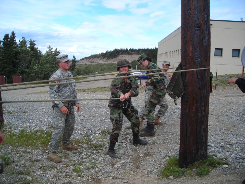 Soldiers in camouflage uniforms pull a rope tied to a wooden pole during an outdoor training exercise
