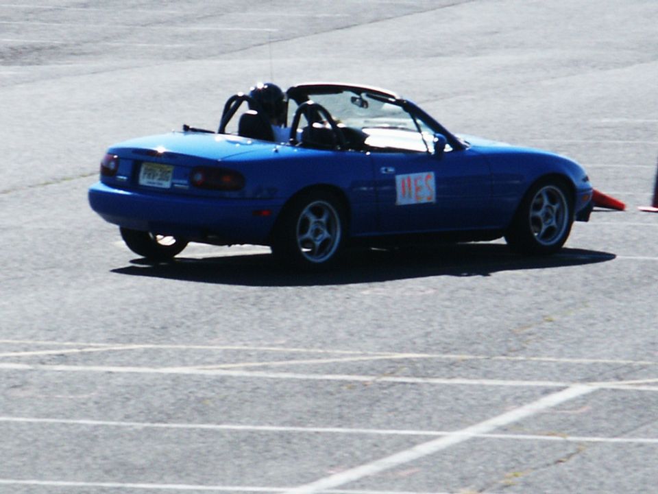 Blue convertible sports car with a roll bar and a driver wearing a helmet, on a paved track