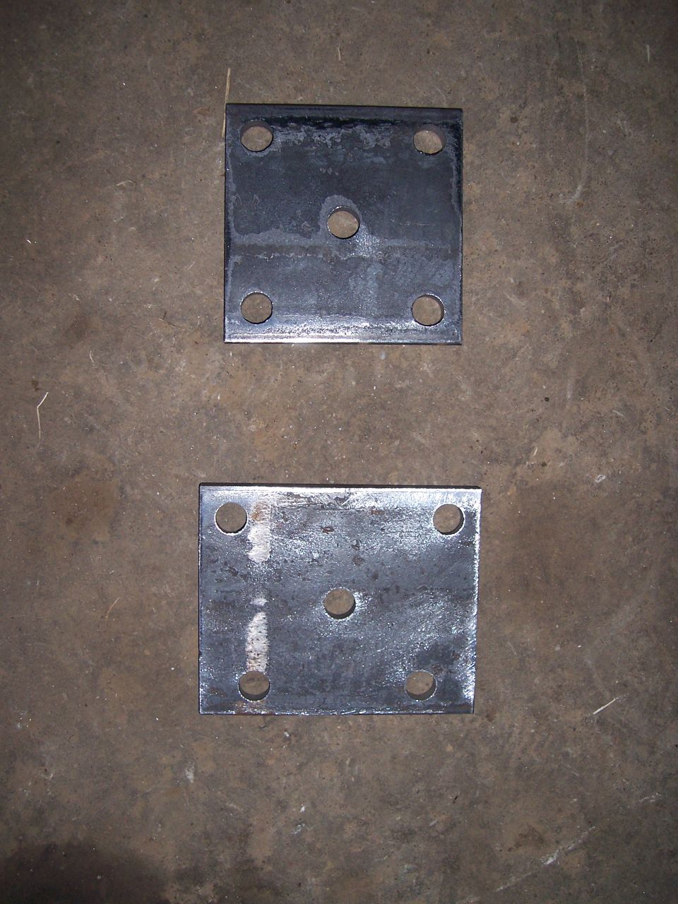 Two square metal plates with five bolt holes each, lying on a concrete floor