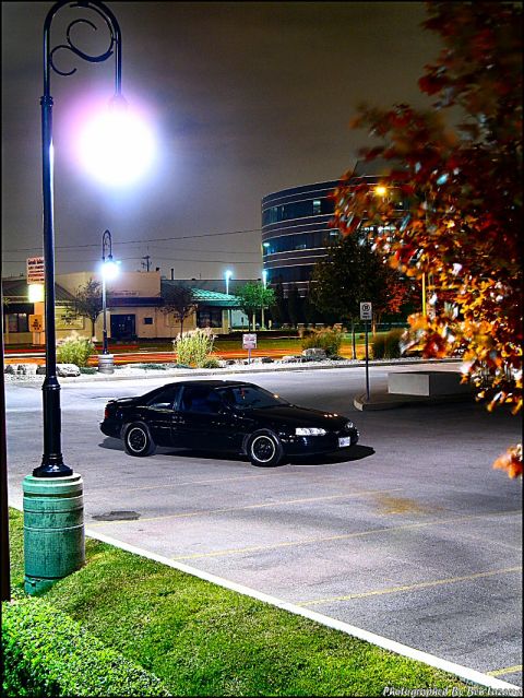 1997 Ford Thunderbird - A black coupe is parked in a lot at night, illuminated by a street lamp and building lights.