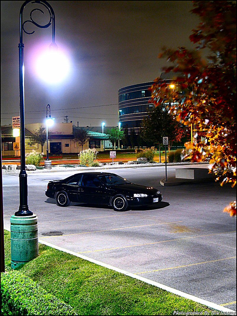 Black coupe parked at night, 3/4 front view. Visible details include a rounded body, distinct headlights, and white-ringed wheels