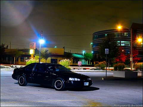1997 Ford Thunderbird - A black coupe is parked in a lot at night, illuminated by streetlights and building lights.