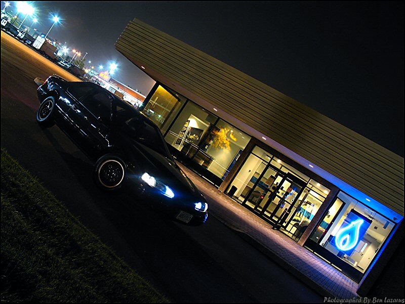 Black coupe with headlights on, parked in front of a building with glass doors and windows at night