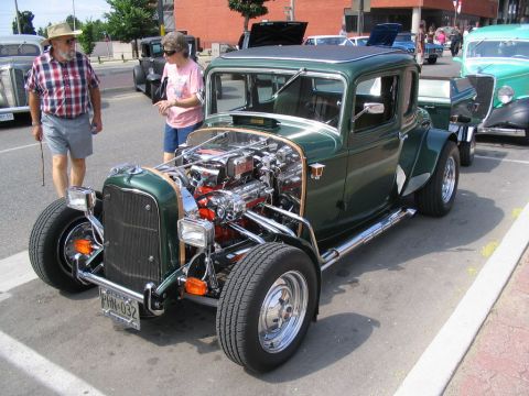 1993 Ford Taurus - A custom green hot rod coupe with a prominent V8 engine is displayed at a car show, with other vehicles and attendees in the background.
