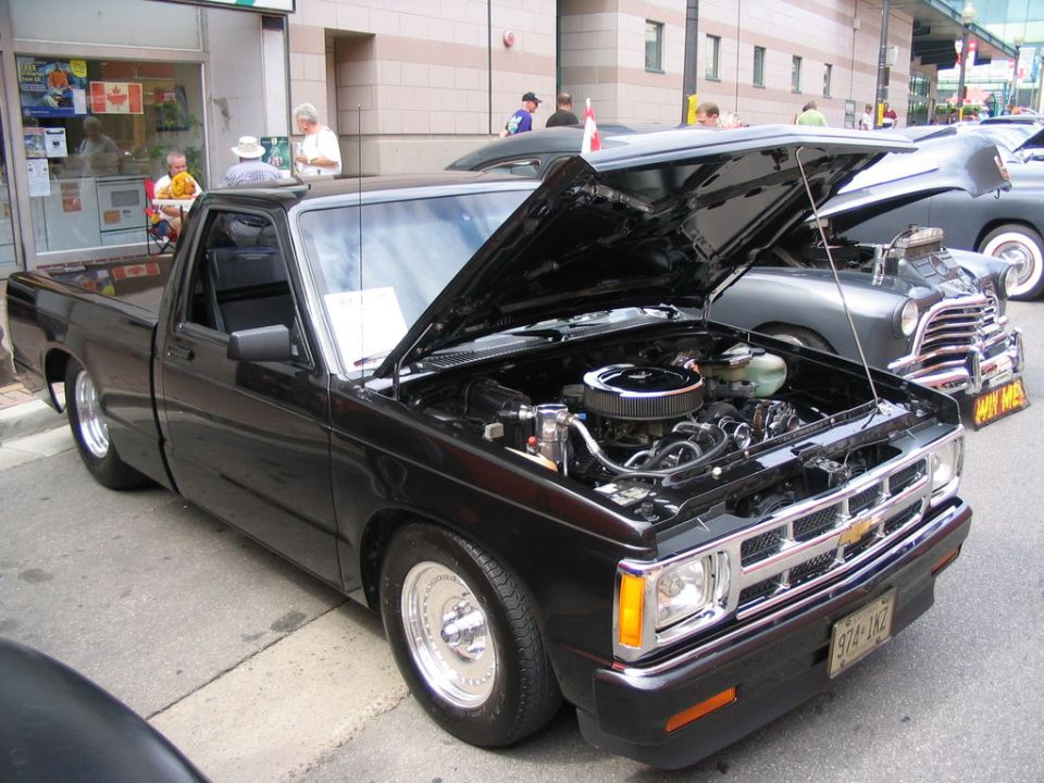 black pickup truck with hood open, showing engine. Another classic car is parked next to it