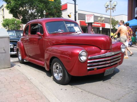 1993 Ford Taurus - A shiny red coupe is parked on the street next to a trash can, with people walking by in the background.