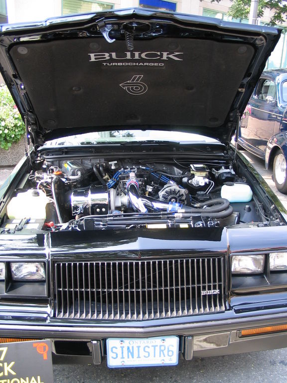 1987 Buick Regal Grand National engine bay with hood open. Turbocharged engine visible. Another car in background