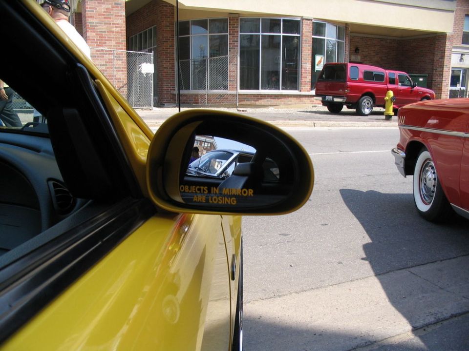 Yellow car's side mirror reflecting a white car interior and "Objects in mirror are losing" text