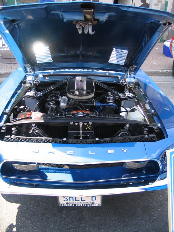 Blue Shelby Mustang, hood open, revealing V8 engine and Shelby badging on grille