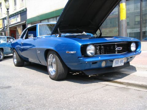 1993 Ford Taurus - A blue 1969 Chevrolet Camaro is parked on a city street with its hood up, revealing the engine compartment.