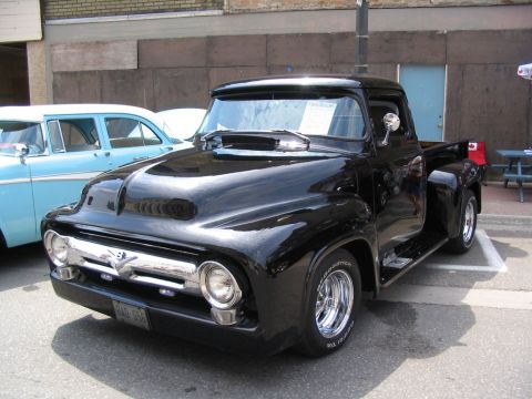 1993 Ford Taurus - A shiny black 1956 Ford F-100 pickup truck is parked on a street next to a light blue sedan.