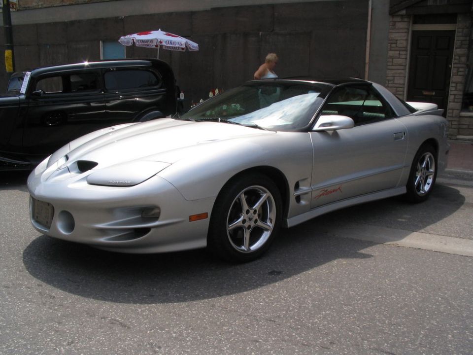 Silver Pontiac Firebird Trans Am with black roof and red side script parked on street, black sedan in background