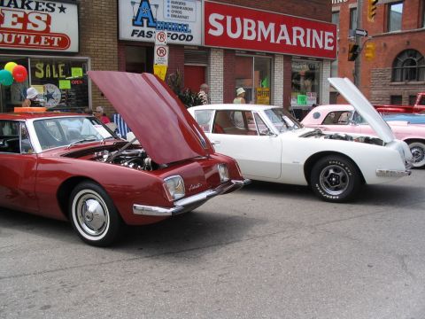 1993 Ford Taurus - Multiple cars with hoods open are displayed at a car show in front of businesses.