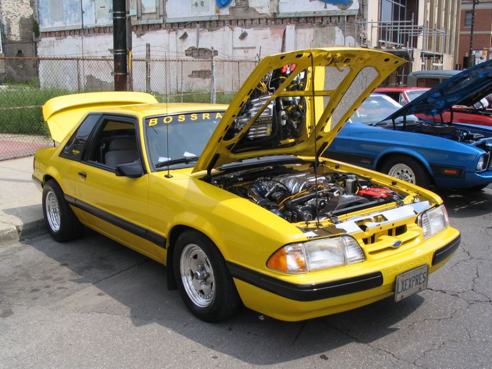 Yellow coupe with hood and trunk open, showing engine. "BOSSRRA" text on windshield. Blue car behind it