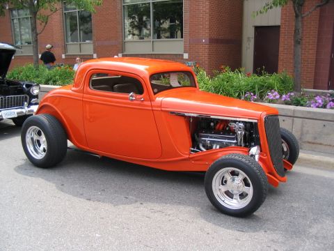 1993 Ford Taurus - An orange hot rod coupe is parked on a street next to a black car, with a brick building in the background.