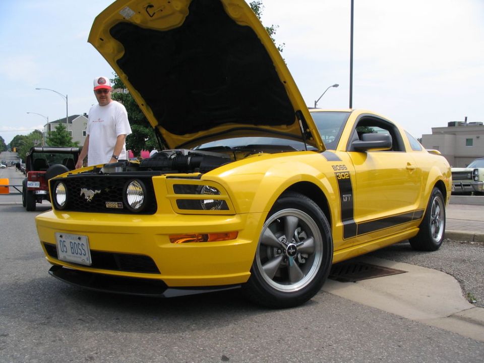 Yellow Ford Mustang with hood open, '05 BOSS' license plate. Man stands nearby. Another vehicle visible in background
