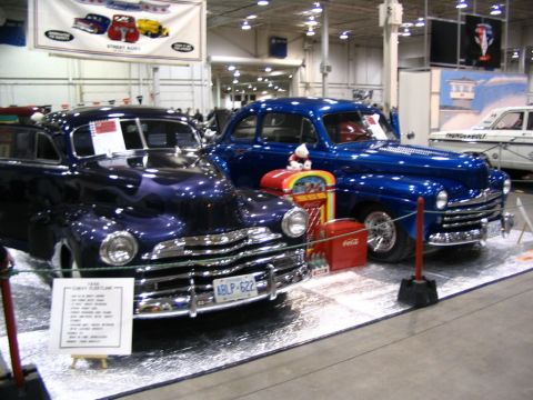 1993 Ford Taurus - Two classic cars, a dark purple and a bright blue sedan, are displayed at an indoor event with a banner overhead.