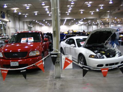1993 Ford Taurus - White coupe and red pickup truck displayed at an indoor event. The coupe's hood is open, revealing the engine.