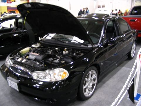 1993 Ford Taurus - Black sedan with hood open, parked indoors next to other vehicles.