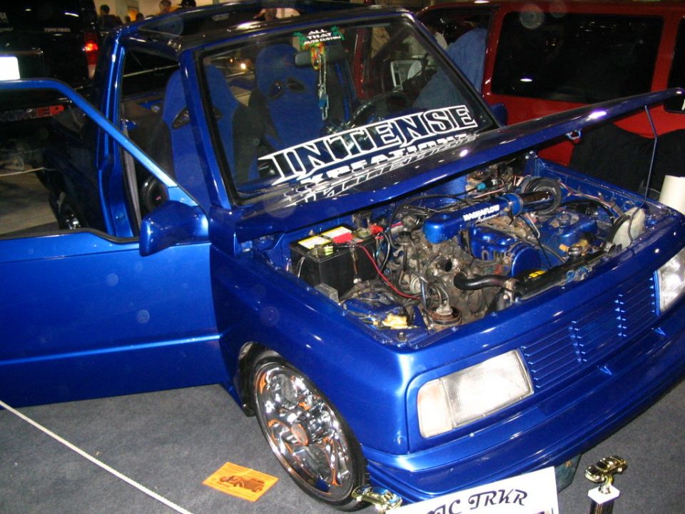 Blue custom pickup truck with aftermarket wheels, racing seats, and "INTENSE" banner on windshield. Engine is visible under the open hood. Another vehicle is partially visible behind it