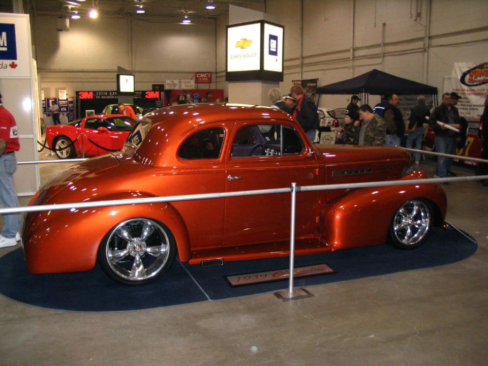 Orange custom hot rod coupe on display, with a red sports car in the background