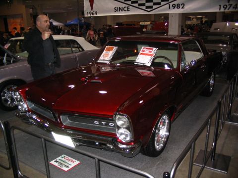 1993 Ford Taurus - A man stands by a shiny red 1965 Pontiac GTO at an indoor car show, with other vehicles visible in the background.