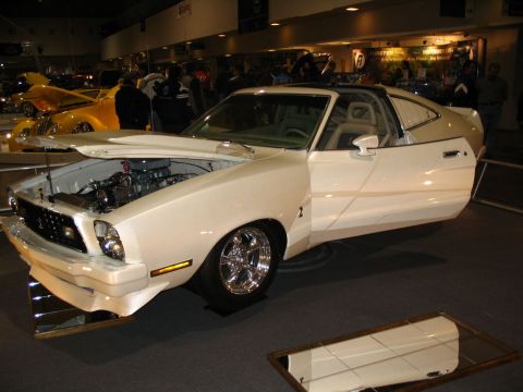 1993 Ford Taurus - A white 1975 Ford Mustang II coupe is displayed with its hood open, revealing the engine. The car features chrome wheels and is part of a l…