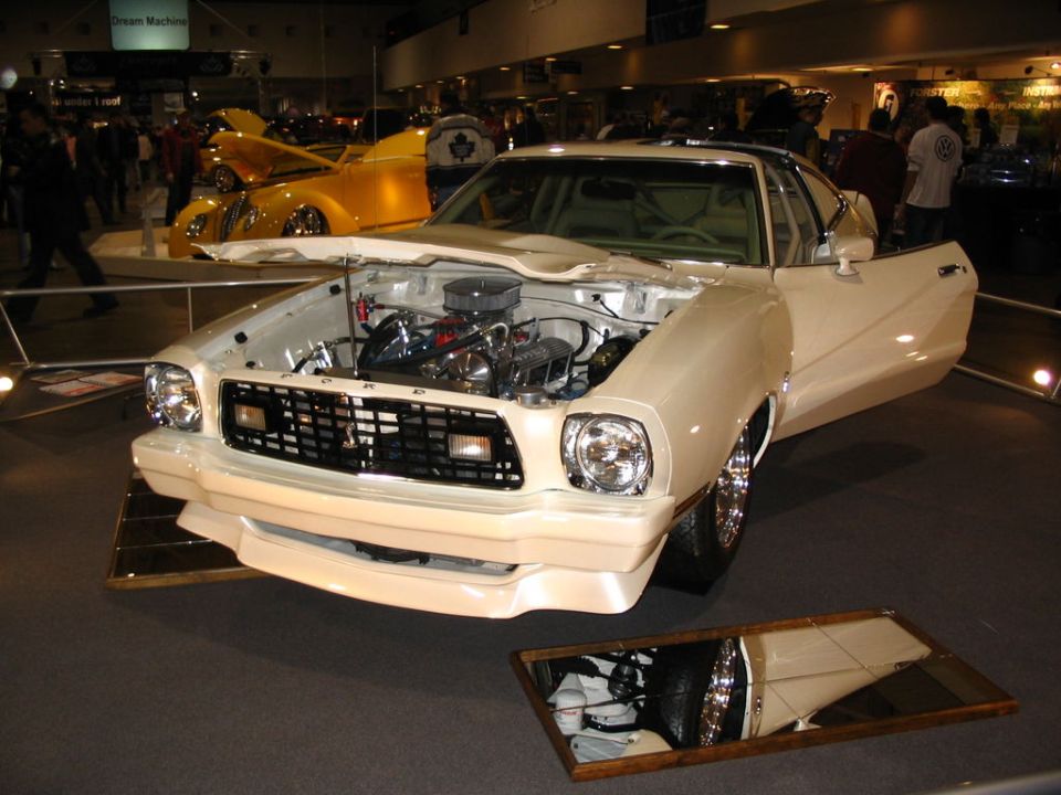 Cream-colored 1975 Ford Mustang II with hood open, showing engine. Yellow car in background