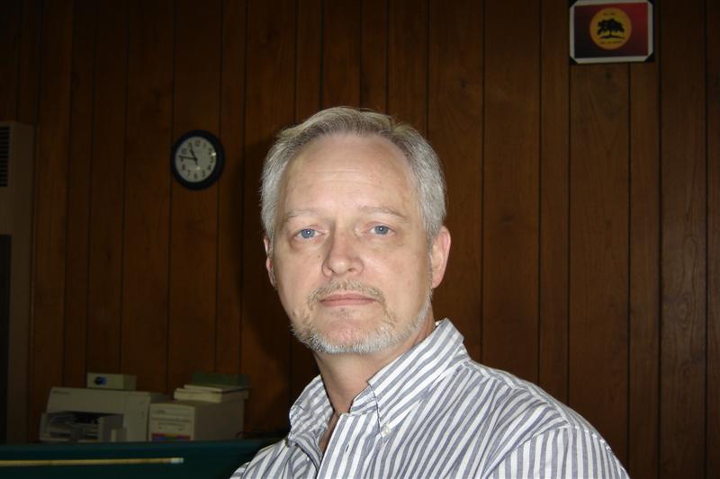 Man with graying hair and goatee wearing a striped shirt. Wood paneled wall behind him with a clock and a framed logo