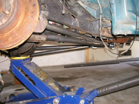 1984 Ford Bronco - The underside of a vehicle is shown, supported by a blue hydraulic jack, with visible suspension and frame components. The website SUPERMOT…