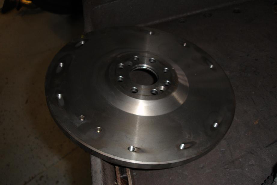 Bare machined steel flywheel with perimeter bolt holes and central hub sitting on a workbench