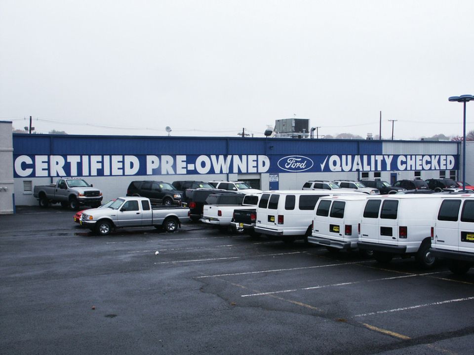 Ford dealership lot with trucks and vans. Building says 'Certified Pre-Owned Ford Quality Checked'