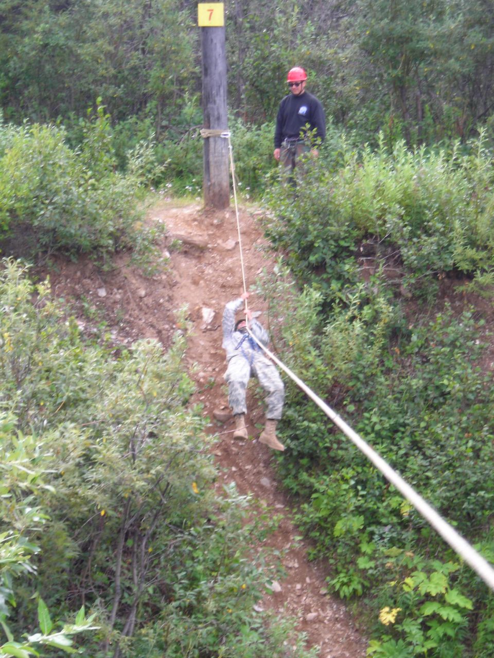 Person in camouflage rappelling down a dirt slope with a rope, a wooden pole with a yellow sign labeled '7' stands nearby, and another person in a red hard hat watches
